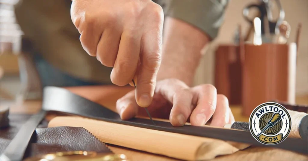 A close-up of a person's hands using an awl tool to carefully punch a hole through a piece of brown leather on a workbench.