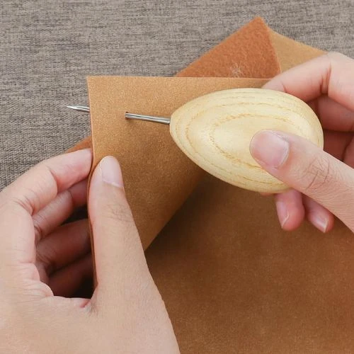 A hand holding the Lineco Wooden Ball Handle Awl, demonstrating its thin metal point piercing a piece of brown book cloth/felt, emphasizing the comfortable grip and extra-heavy duty build.